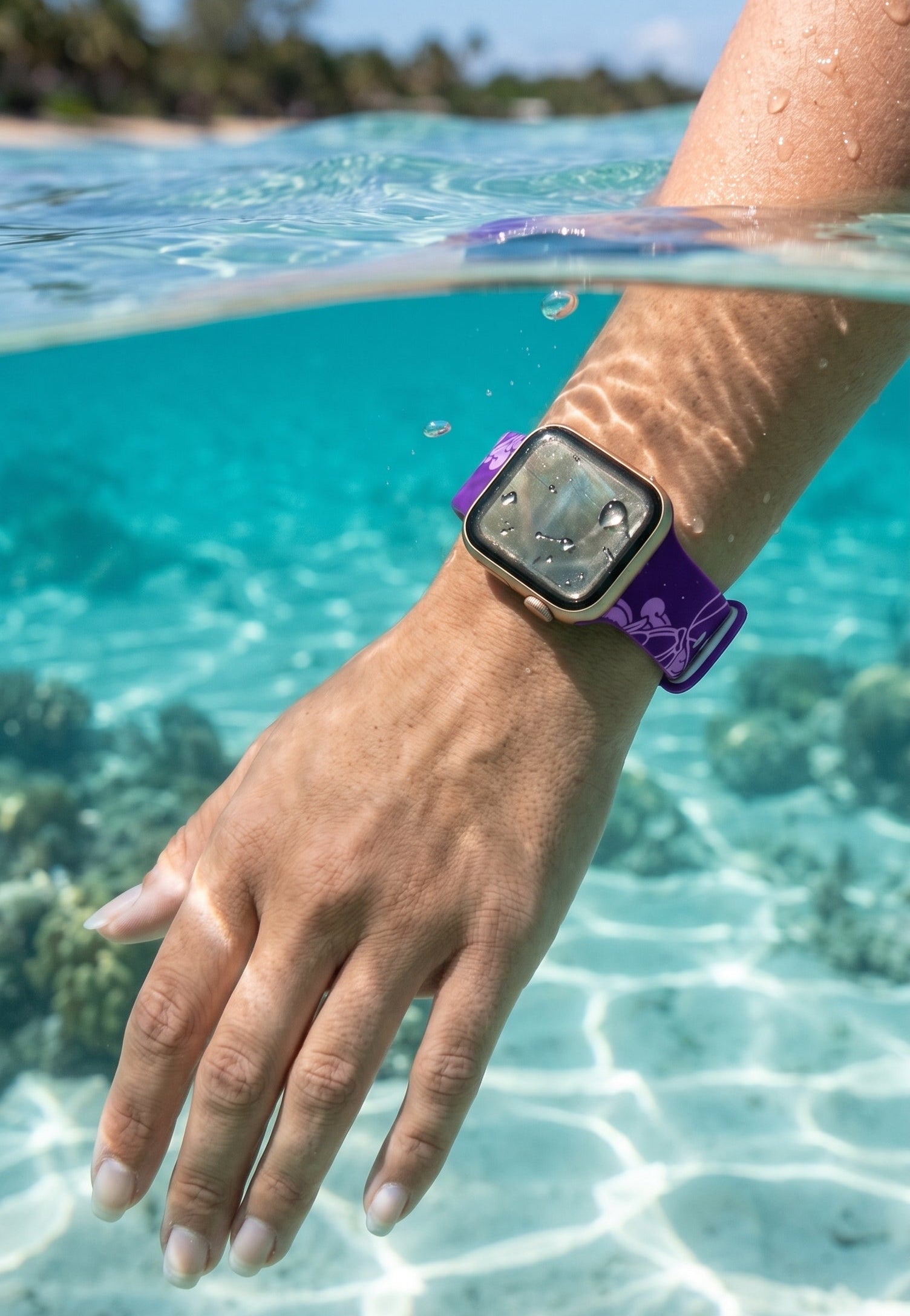 A person's hand and forearm wearing a smartwatch with a purple strap, photographed underwater in clear turquoise ocean water with coral and a beach visible above the surface.